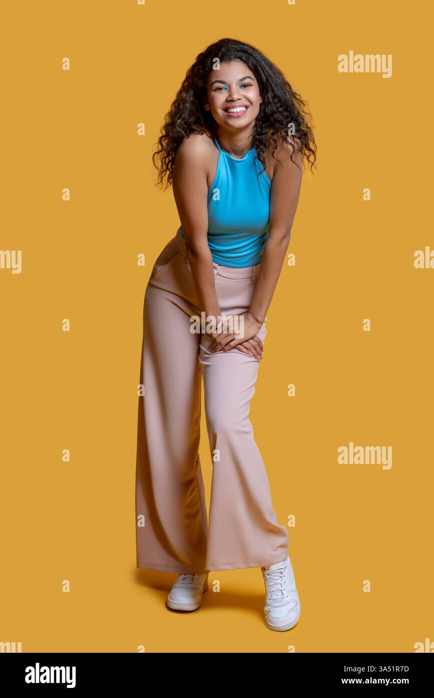 Helles Atelierporträt einer lächelnden Afroamerikanerin, die beiläufig an eine Wand posierte. Ihre entspannte Pose und der fröhliche Ausdruck machen dieses Bild vielseitig für Lifestyle-, Mode- oder Beauty-Kampagnen. Hervorragend geeignet für Branding, das in Marketingmaterialien ansprechbare, positive Bilder sucht. Stockfoto