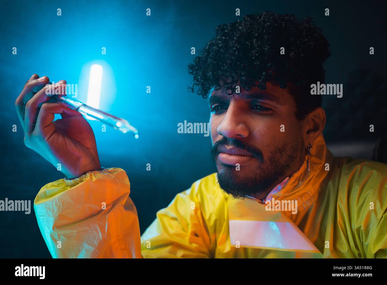Hispanischer Mann in einem Gefahrenanzug inspiziert eine flüssige Chemikalie mit einer Glaspipette in einer dunklen Laborumgebung. Diese professionelle Chemieszene beleuchtet Laborarbeiten und wissenschaftliche Untersuchungen. Ideal für Themen wie Laborsicherheit, chemische Forschung und Industriewissenschaft. Stockfoto