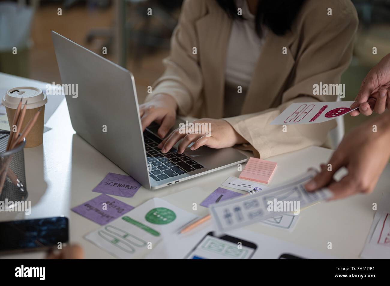 Professionelle asiatische Grafikdesignerin arbeitet in einem modernen Büro mit ihrem Team zusammen und arbeitet während eines Design-Meetings an einem Laptop. Das Bild betont Zusammenarbeit, Kreativität und UX-UI-Planung in einem modernen Arbeitsplatz. Ideal für Designstudios, Technikteams und Marketingkampagnen. Stockfoto