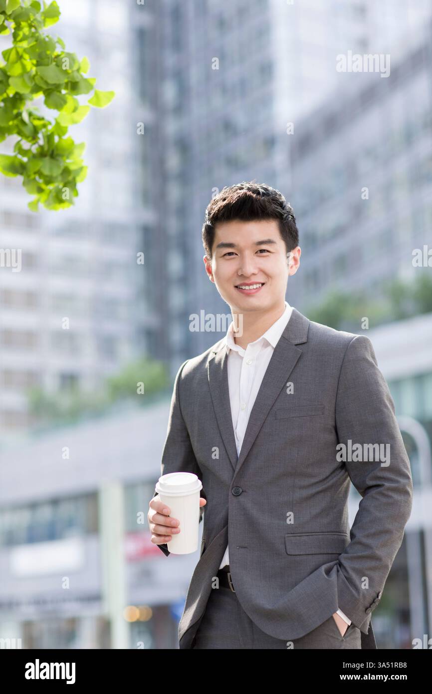 Chinesischer Geschäftsmann mit Kaffeetasse, der an sonnigen Tagen in der Nähe von Gebäuden in der Stadt steht Stockfoto