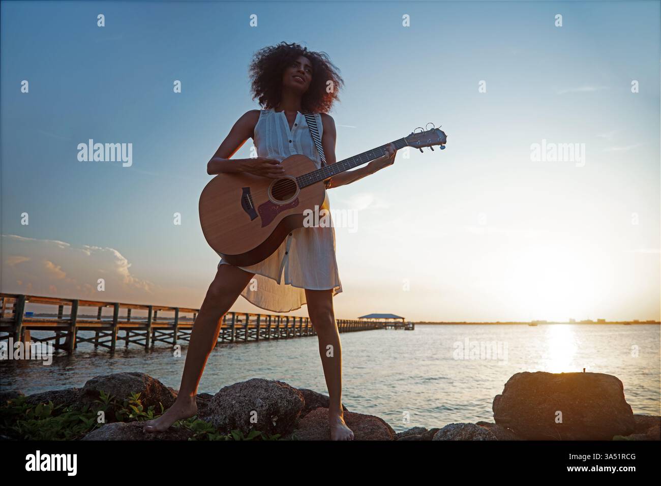 Sorglose junge afroamerikanische Frau, die sich im Sommerurlaub entspannt und bei Sonnenaufgang Gitarre am Meer spielt. Eine entspannte Küstenszene, die Reisen, Musik und Ruhe am frühen Morgen verbindet. Ideal für Strandurlaube, Freizeit und musikalische Momente am Ufer. Stockfoto