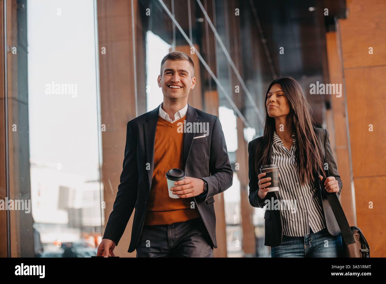 Fröhliche hispanische Geschäftsfrau und kaukasischer Geschäftsmann reisen auf Geschäftsreise, Kaffee in der Hand, schlendern mit Gepäck durch einen Flughafen. Ideal für Geschäftsreisen, Teamarbeit und internationale Geschäftsthemen. Stockfoto
