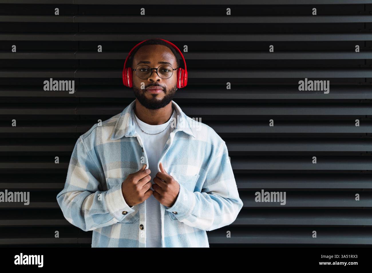 Serious Black man trägt Brille und Kopfhörer, steht in einer urbanen Straßenszene vor einer strukturierten schwarzen Wand. Ideal für Lifestyle-, Musik- und technologieorientierte Kampagnen. Geeignet für professionelle Profile und Materialien für urbane Mode. Stockfoto