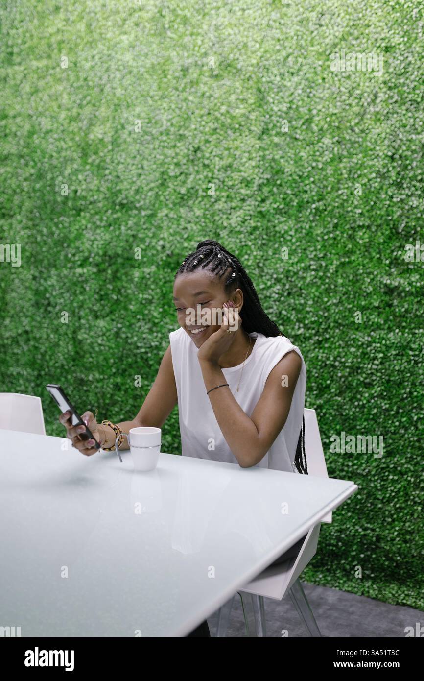 Afroamerikanerin sitzt drinnen und legt ihr Kinn auf ihre Hand, während sie ein Smartphone durchsucht. Dieses zeitgenössische Lifestyle-Bild passt zu Themen wie Technik, soziale Medien und Home Office. Perfekt für Konnektivität, Online-Dienste und entspannte Alltagsmomente. Stockfoto