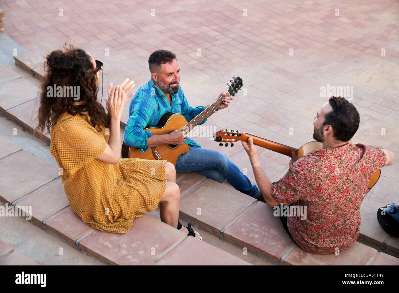 Aus einem erhöhten Blickwinkel sitzt eine Gruppe von Musikern auf Stufen mit Akustikgitarren, spielt und lacht. Eine fröhliche Frau tritt in die Szene ein und schafft einen warmen, zwanglosen Musikmoment draußen in der Stadt. Geeignet für Lifestyle, Straßenauftritte und Hobbyinhalte. Stockfoto