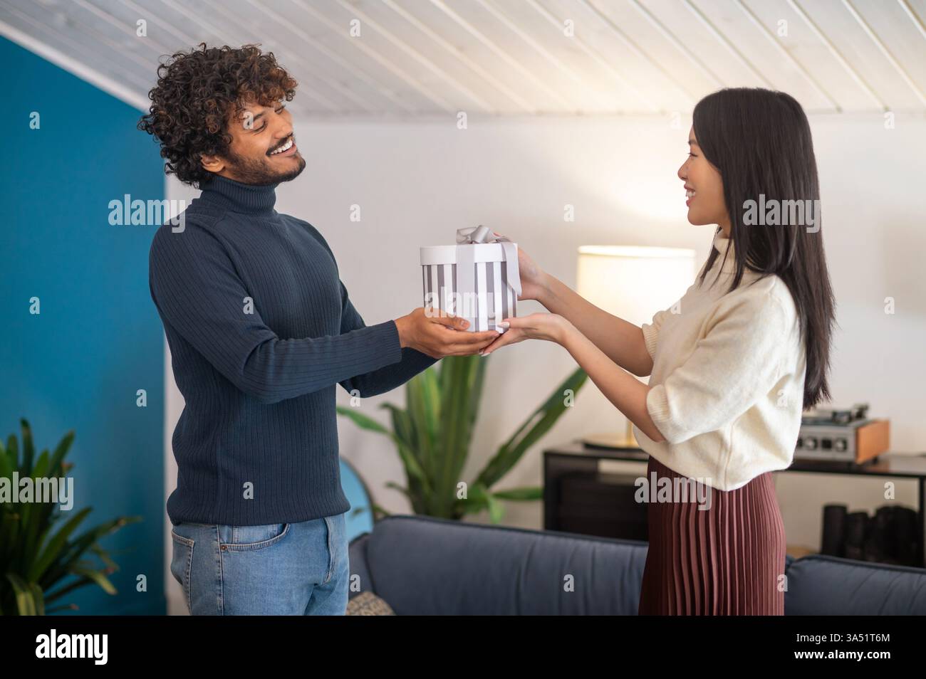 Fröhlicher Urlaub zu Hause, als ein asiatischer Mann einer lächelnden hispanischen Frau ein Geschenk schenkt. Die Szene ist drinnen mit warmer Beleuchtung, die den Geschenketausch und die festliche Feier betont. Ein vielseitiges Bild für Weihnachten, Familienanlässe und vielfältige Beziehungen. Stockfoto