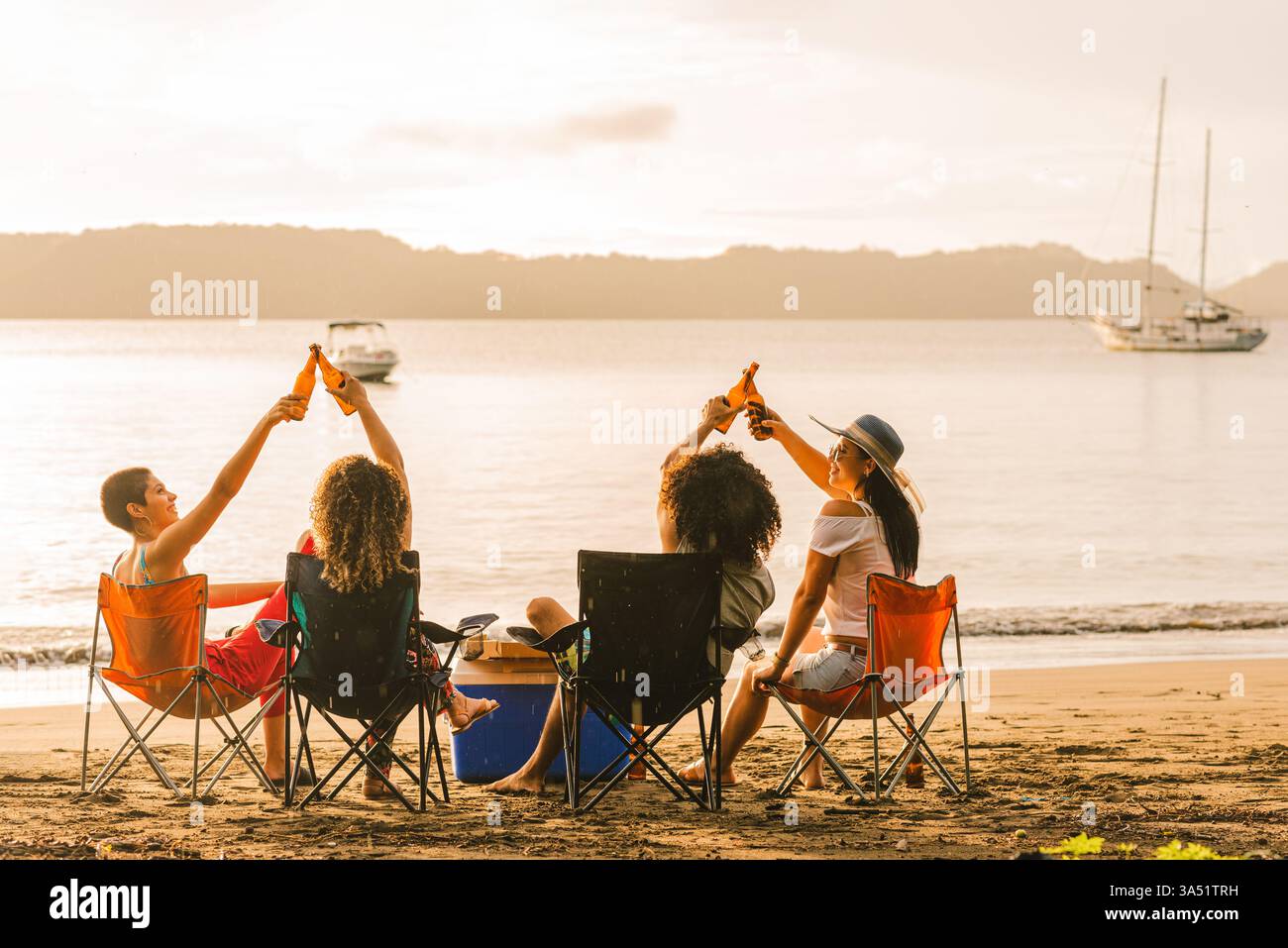 Fröhliche Gruppe junger Freunde an einem Sandstrand teilt sich einen Toast und lacht. Sie sitzen auf Stühlen und klopfen Bierflaschen in einem sorgenfreien Sommermoment an. Ein vielseitiges Lifestyle-Image, perfekt für Reisen, Freundschaften und Outdoor-Feiern. Stockfoto