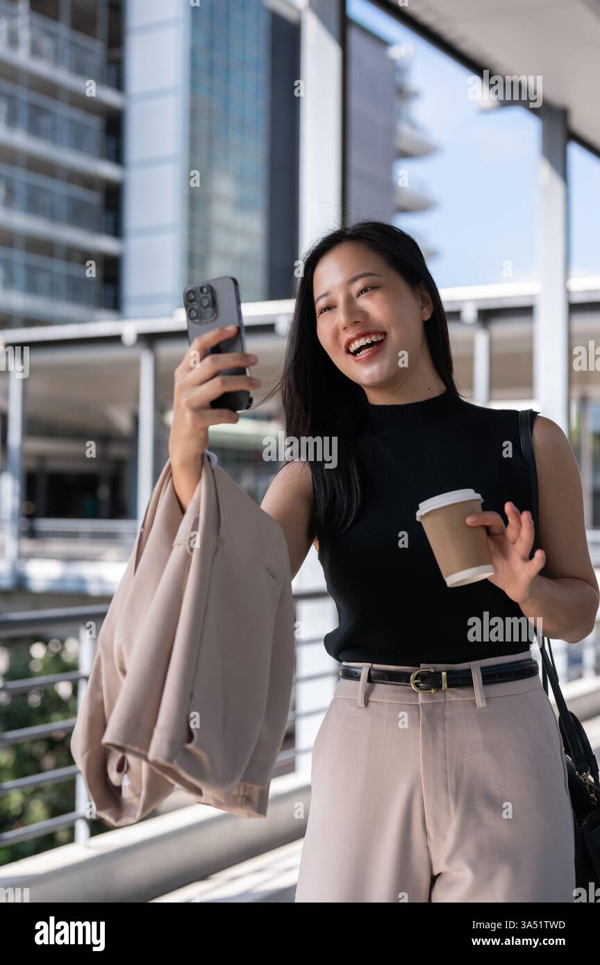 Lächelnde asiatische Frau mit schwarzem ärmellosem Oberteil, die Videoanrufe führt, während sie eine Tasse Kaffee hält und in der Stadt auf einem Fußgängersteg steht Stockfoto