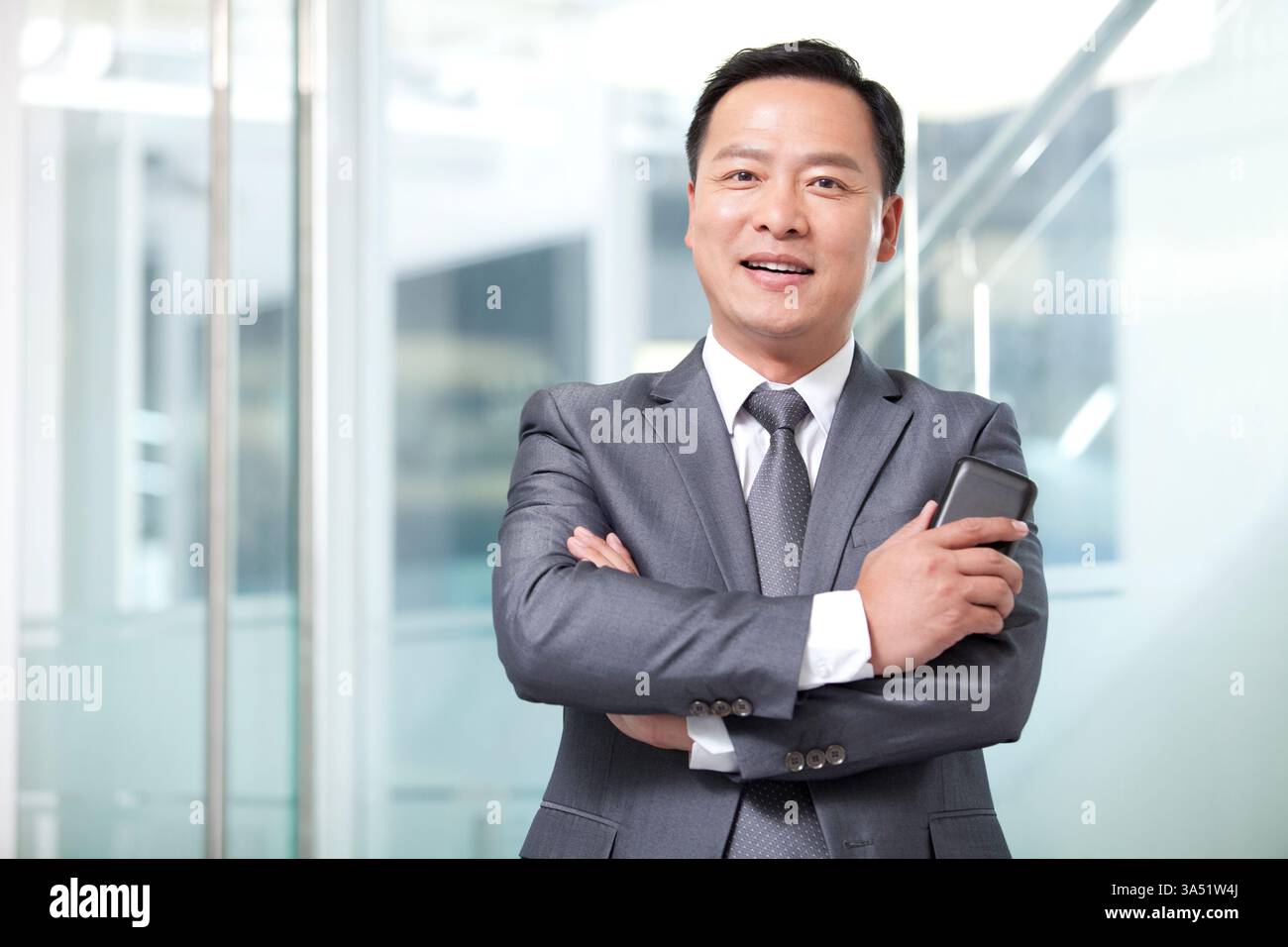 Lächelnder chinesischer Geschäftsmann mittleren Alters, der mit überkreuzten Armen im Amt steht Stockfoto