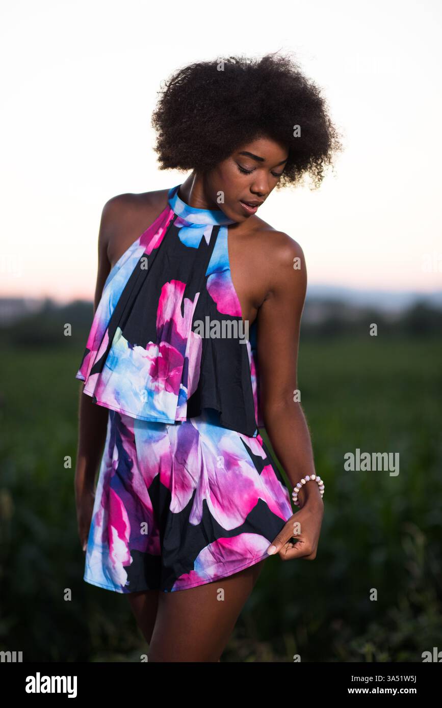 Porträt einer lächelnden Afroamerikanerin in einem Sommerkleid an einem warmen Abend. Ein stilvolles Lifestyle-Bild, das natürliches Make-up und lockiges Haar für Mode- und Schönheitsmerkmale aufnimmt. Ideal für Mode-, Lifestyle- und Marketingkampagnen. Stockfoto