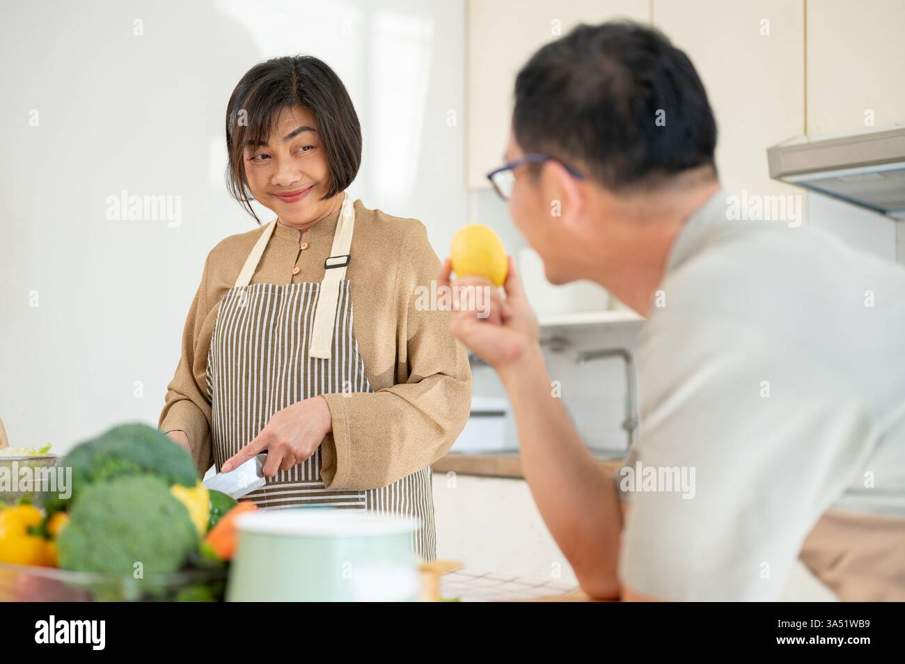 Ein glücklicher asiatischer Ehemann plaudert mit seiner Frau, während sie in der Küche das Frühstück kocht. Der Moment hebt das häusliche Leben, die Wärme und die familiäre Bindung hervor. Ideal für Lifestyle, Beziehungen und Hausmannskost. Stockfoto