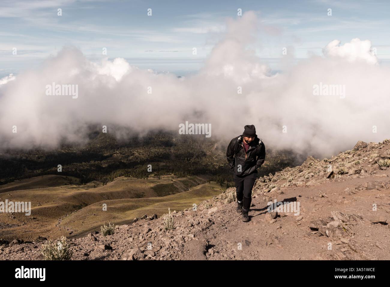 Ein Mann, der auf einem felsigen Bergpfad hinunterblickt, mit einer riesigen Landschaft aus Wolken und Hügeln im Hintergrund. Dramatische Naturlandschaften, die Abenteuer, Natur und Fitness für Reisen oder Wanderungen einfangen. Stockfoto