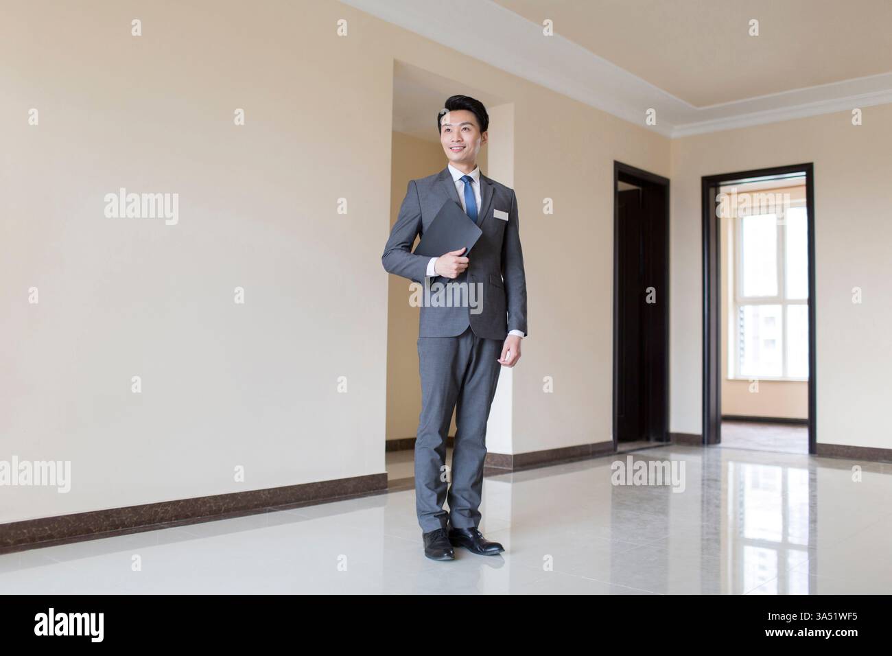 In einem modernen Haus in Peking hält ein lächelnder chinesischer Immobilienmakler in einem grauen Anzug ein Klemmbrett. Dieses Image ist ideal für Immobilienverkäufe, Immobiliendienstleistungen und Wohnungseigentum-Kampagnen in China. Stockfoto