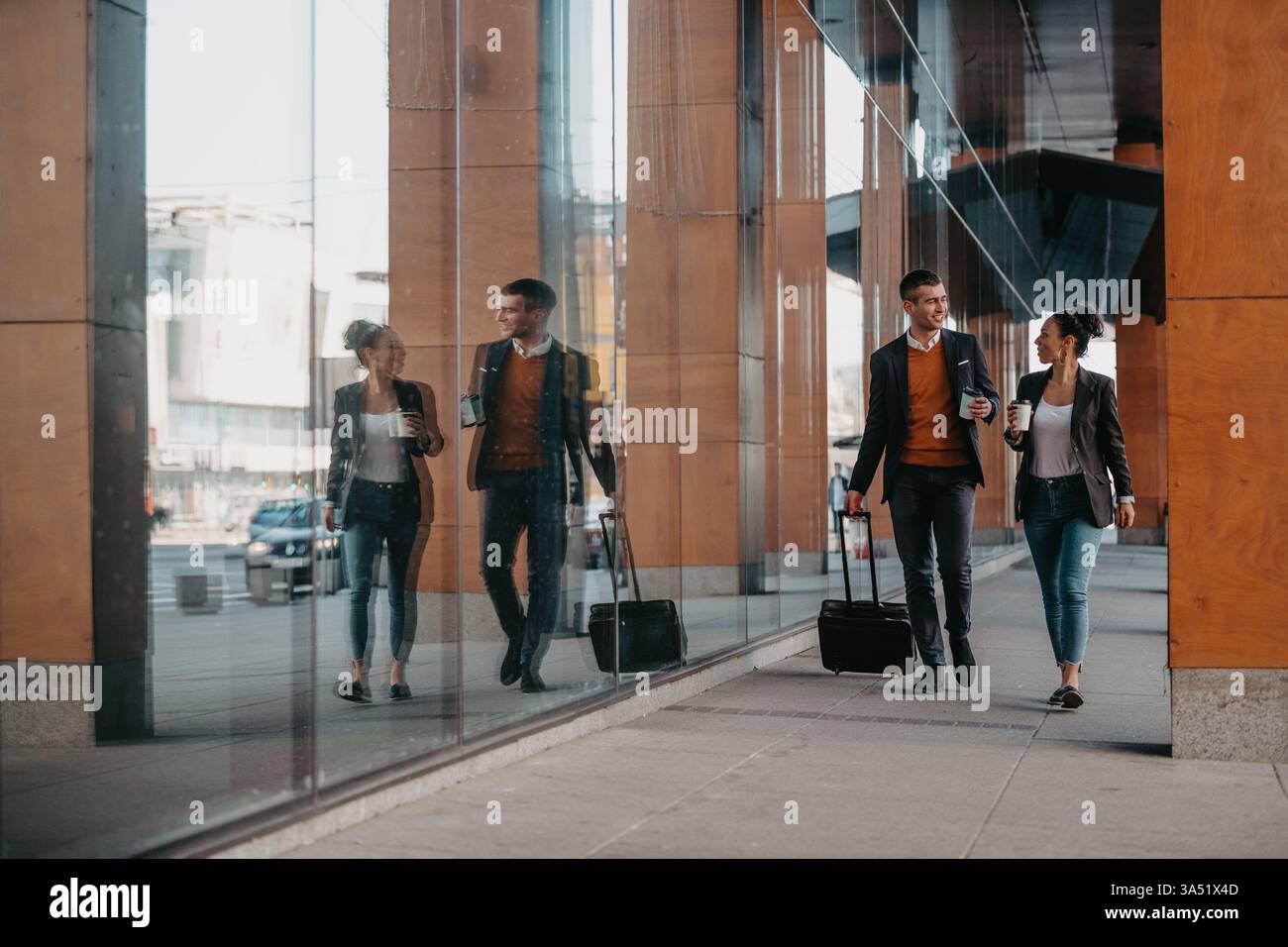 Fröhliche hispanische Geschäftsfrau und kaukasischer Geschäftsmann reisen zusammen auf einer Geschäftsreise und plaudern bei Kaffee, während sie mit dem Gepäck spazieren gehen. Lebendige urbane Szene, geeignet für Geschäftsreisen, internationale Geschäfte und Networking-Themen. Hervorragend geeignet für multiethnische Geschäftskooperationen und moderne Reisekampagnen. Stockfoto