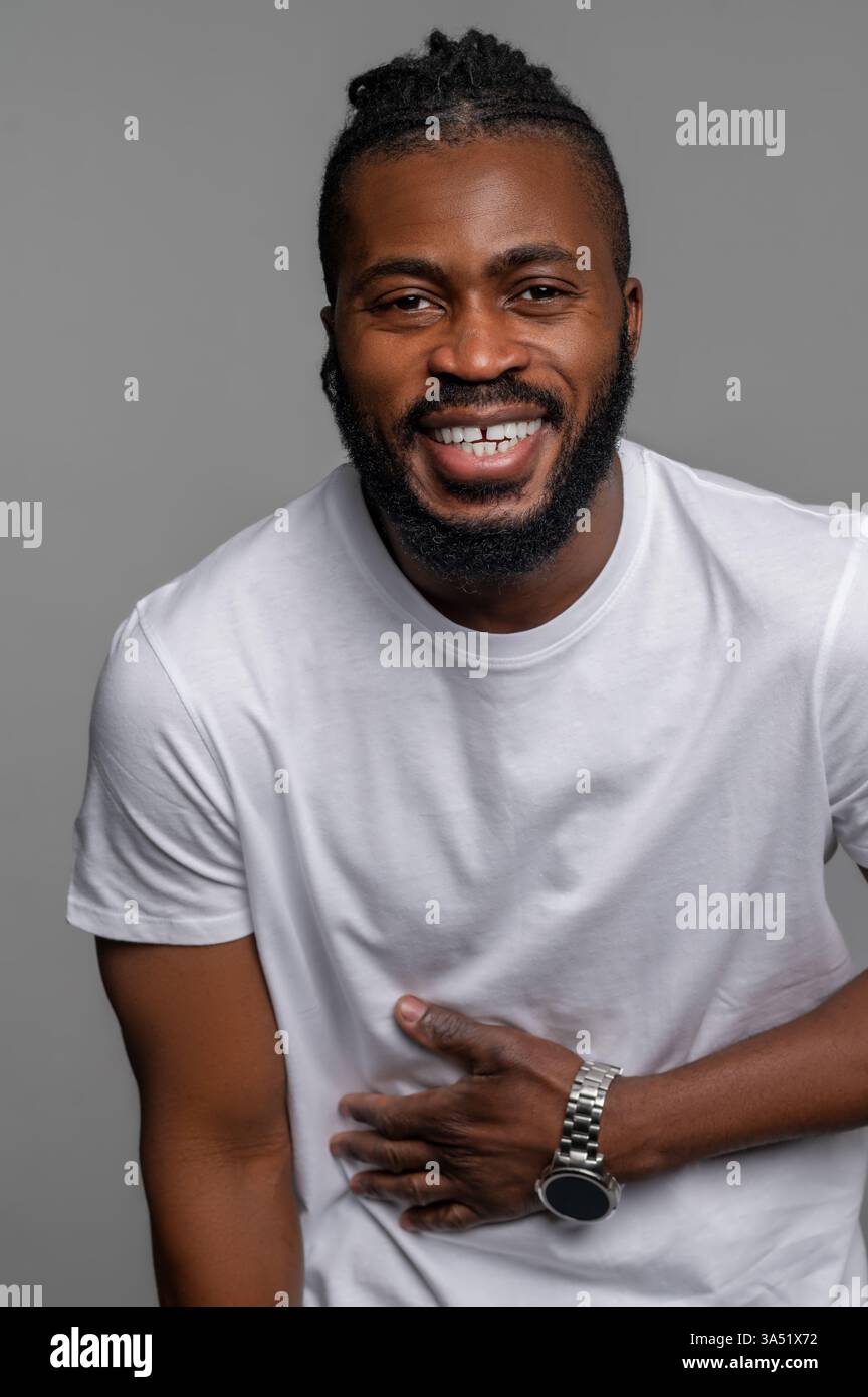 Warmes Porträt eines Afroamerikaners, der lacht und seinen Bauch hält. Dieses Bild vermittelt eine fröhliche, positive Stimmung, die für Lifestyle- und Marketingkontexte geeignet ist. Das Motiv trägt ein lässiges Outfit und wird in einem Studio im Innenbereich fotografiert, um einen ansprechbaren, ansprechbaren Look zu kreieren. Stockfoto