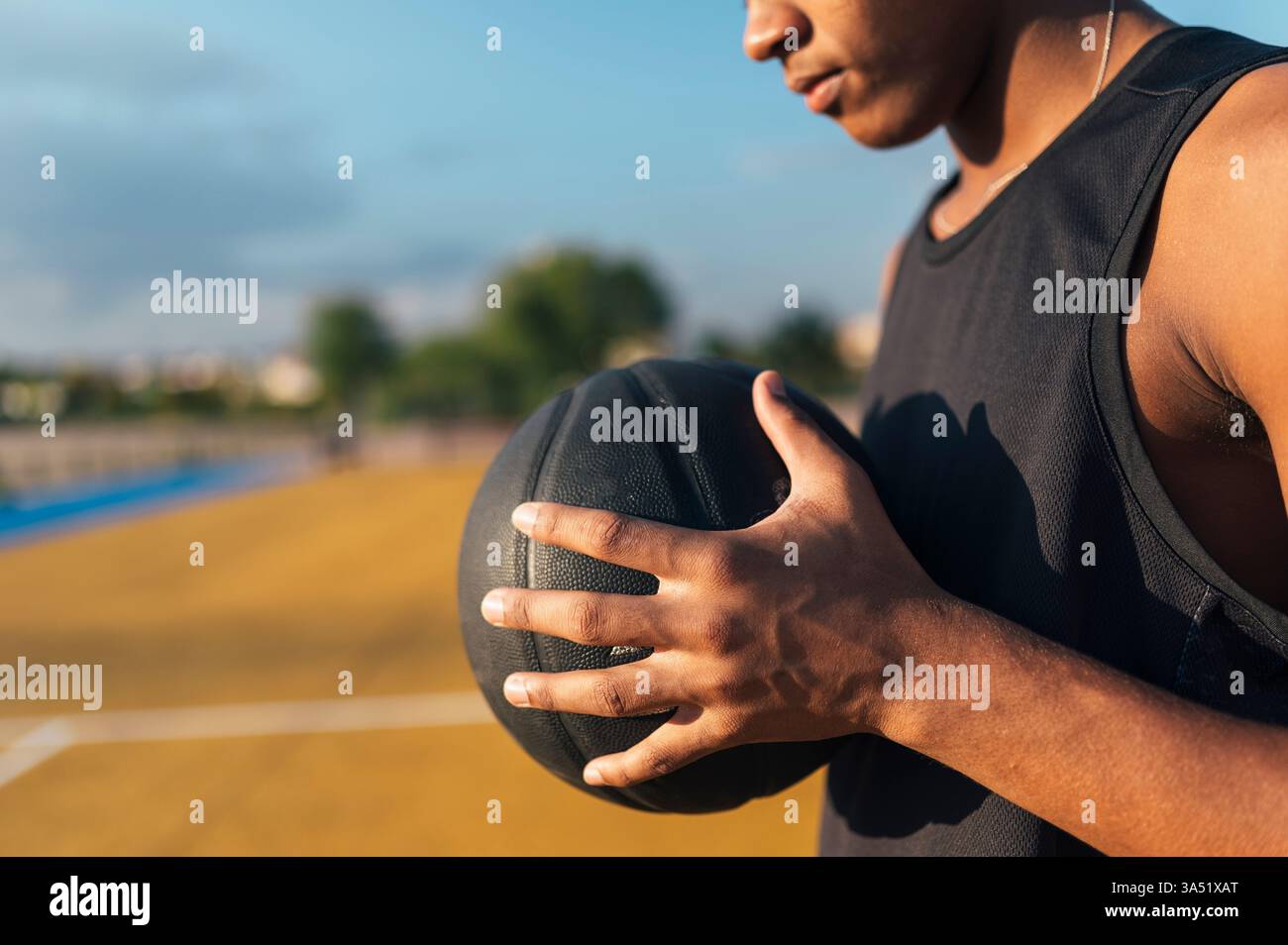 Nicht wiedererkennbarer afroamerikanischer Basketballspieler in Uniform mit Ball auf einem hellen Spielplatz im Freien. Die zugeschnittene Aufnahme lässt Platz für Text und betont Action und sportliche Energie. Geeignet für Basketball, Sportmarketing und Jugendwettbewerbe. Stockfoto