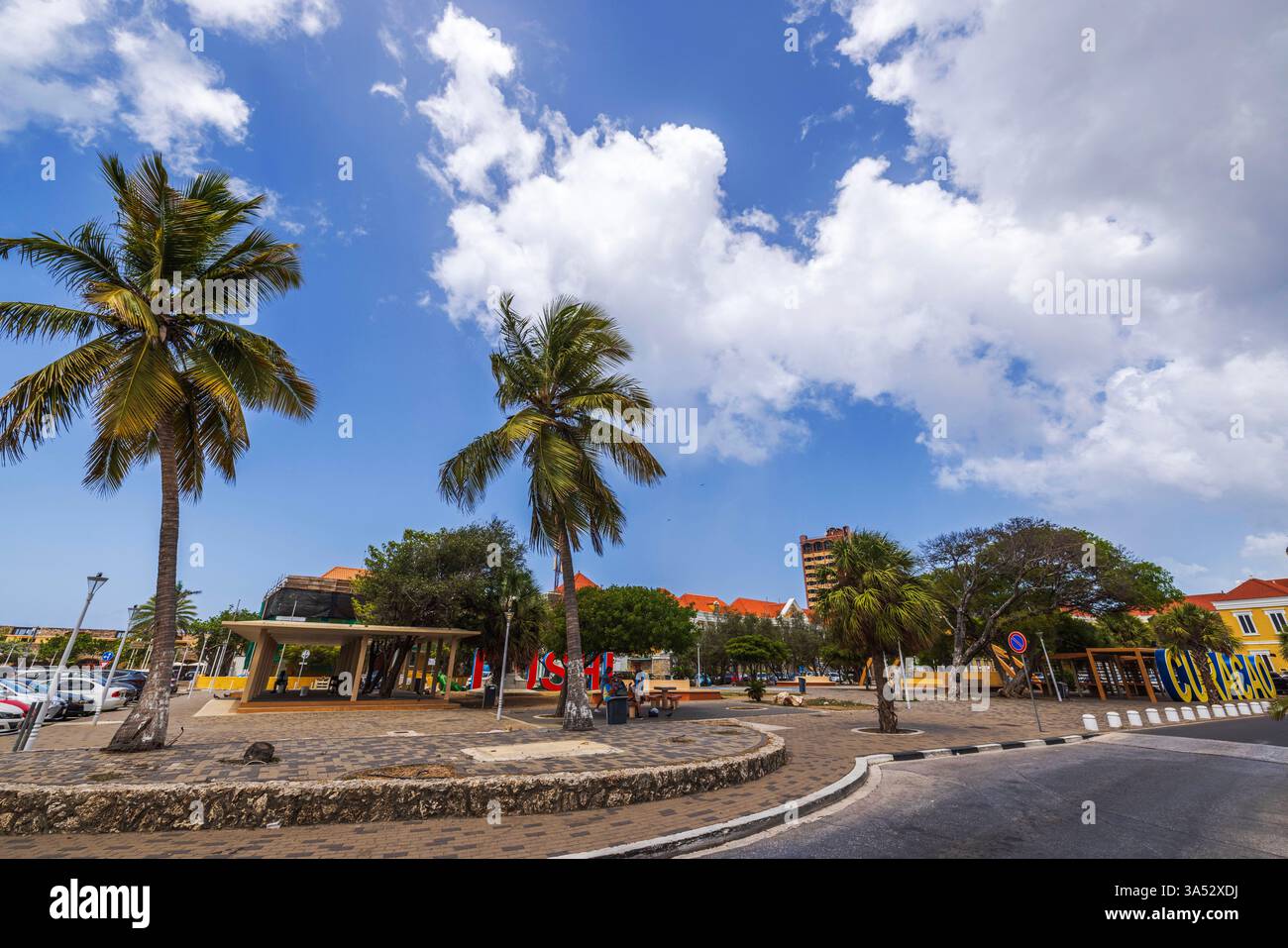 Willemstad-Platz mit Curacao-Schild zwischen Palmen und farbenfrohen Gebäuden vor blauem Himmel mit weißen Wolken. Willemstad. Curacao. Stockfoto