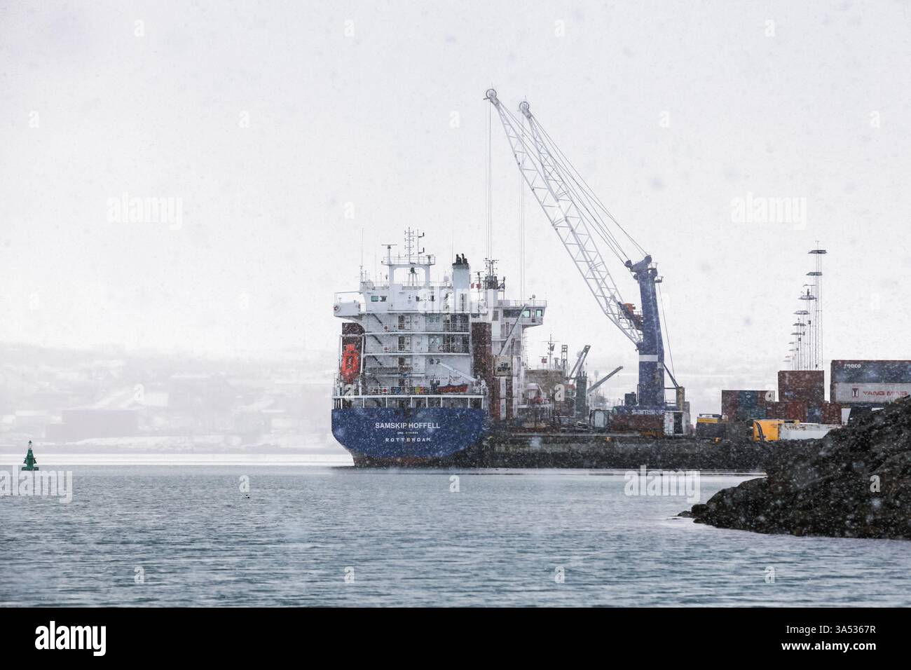 Reykjavik, Island - 4. April 2017: Großes Frachtschiff an einem belebten Hafen, umgeben von Containern und Kränen, bei Schneefall. Spiegelt Mariti wider Stockfoto