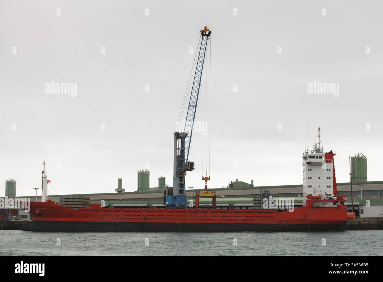 Hafnarfjordur, Island - 4. April 2017: Ein rotes Frachtschiff wird in einem Industriehafen angedockt und verfügt über einen Kran, der Güter und nahegelegene Einrichtungen entlädt. Stockfoto