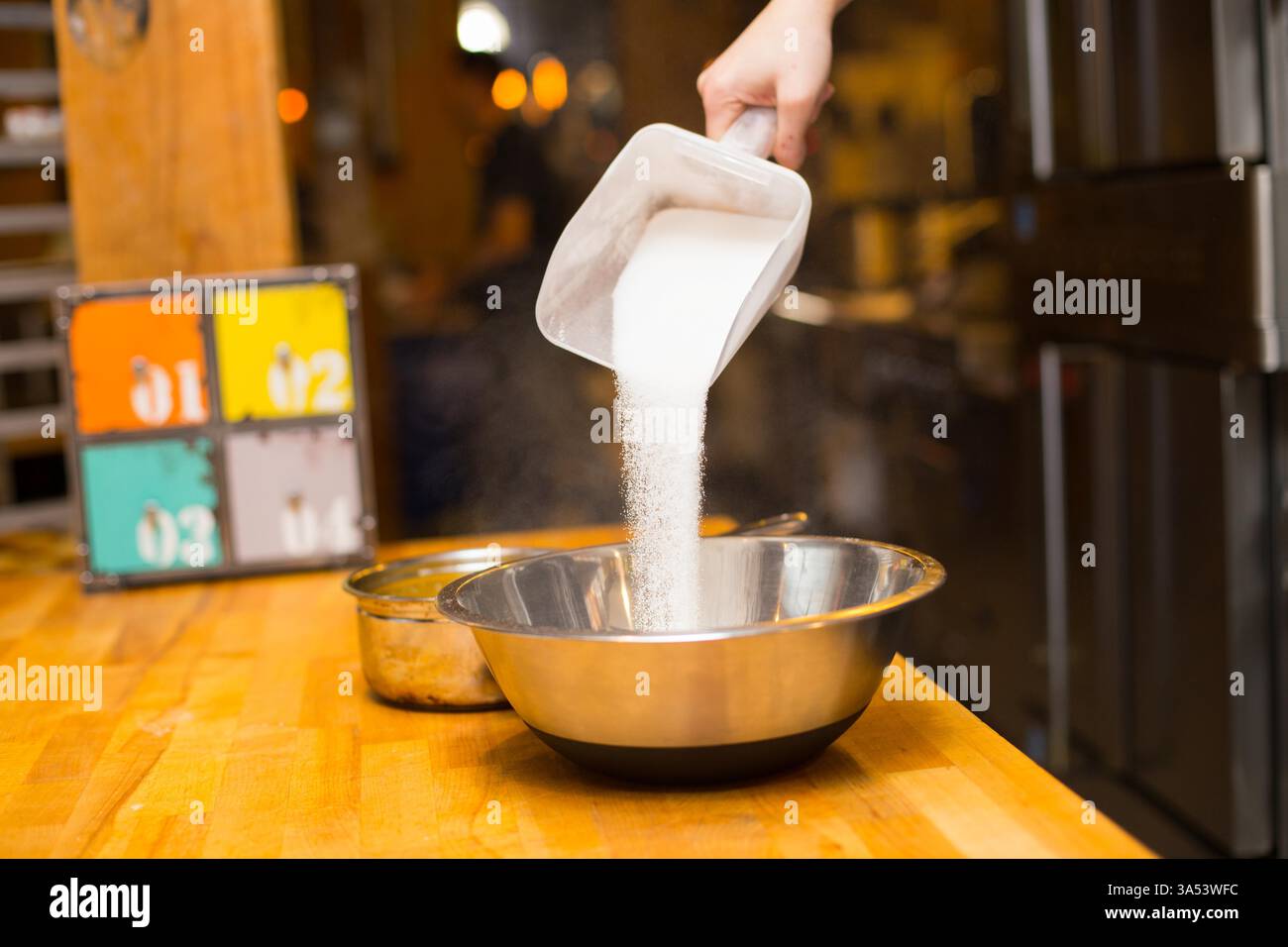 Aus einem Kunststofflöffel Zucker in eine Edelstahlschale gießen Stockfoto