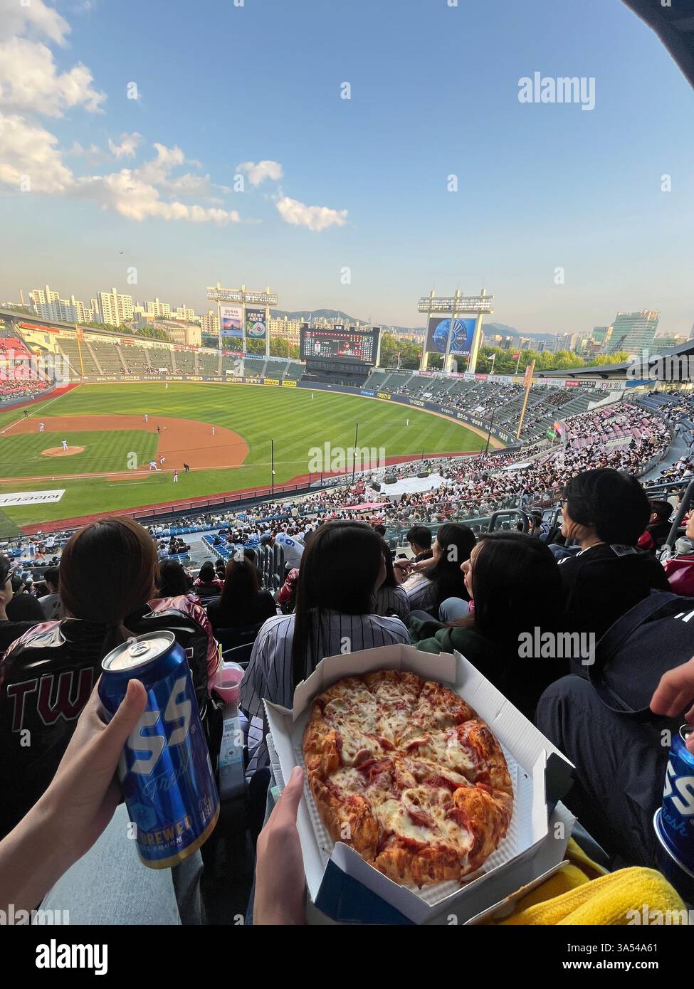 Eine Person genießt Pizza und Bier, während sie ein professionelles Baseballspiel in einem überfüllten Stadion am Abend ansieht. - Smartphone-aufgenommenes Stockfoto