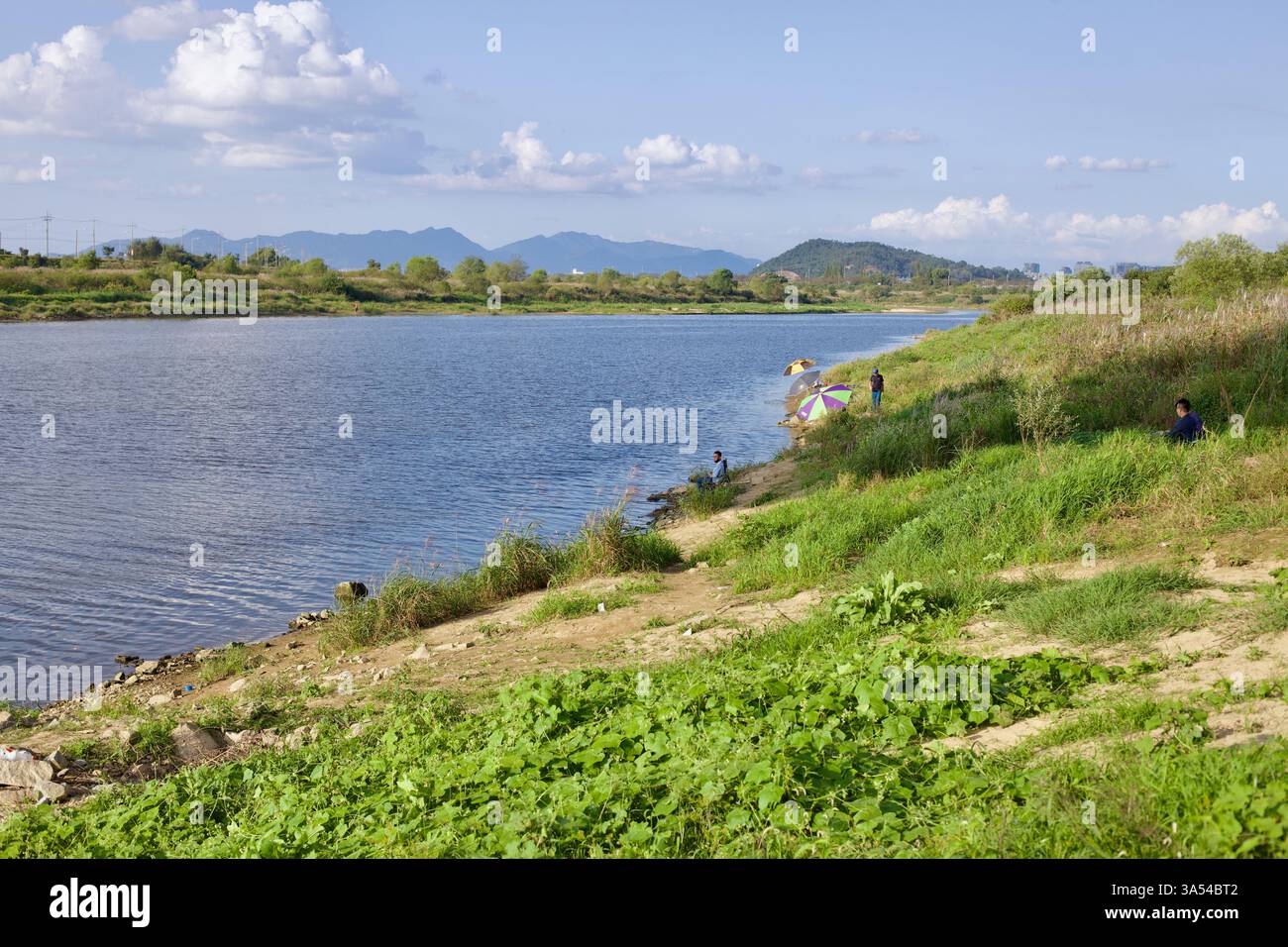 Gwangju, Südkorea - 24. September 2020: Menschen fischen am Ufer des Yeongsangang River, beschattet von bunten Regenschirmen, mit sanften Hügeln und Stockfoto
