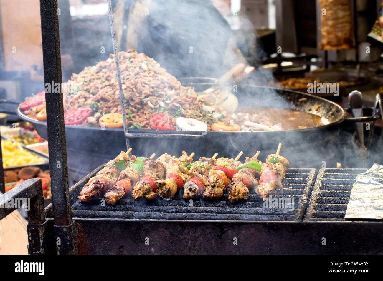 Fleisch- und Gemüsespieße, die auf einem Straßenmarkt über rauchigen Flammen grillen, mit einer großen Paella-Pfanne im Hintergrund. Lebhafte Street Food Atmosphäre Stockfoto