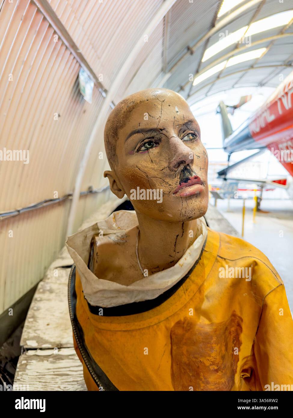 Getragener Schaufensterpuppe-Kopf in orangefarbenem Anzug im Flugzeughangar mit einem alten Militärjet im Hintergrund - Smartphone-aufgenommenes Stockfoto
