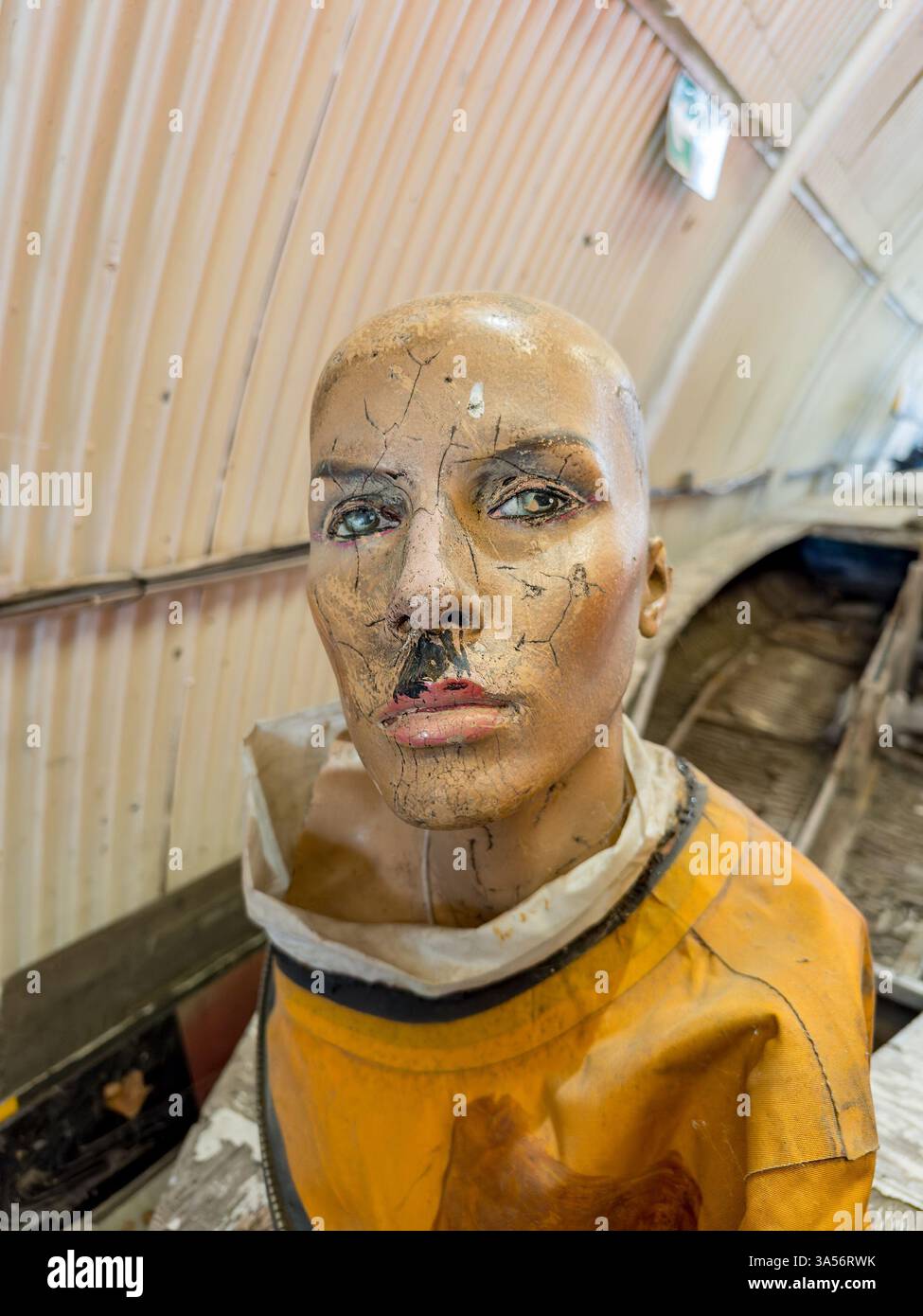 Getragener Schaufensterpuppe-Kopf in orangefarbenem Anzug im Flugzeughangar mit einem alten Militärjet im Hintergrund - Smartphone-aufgenommenes Stockfoto