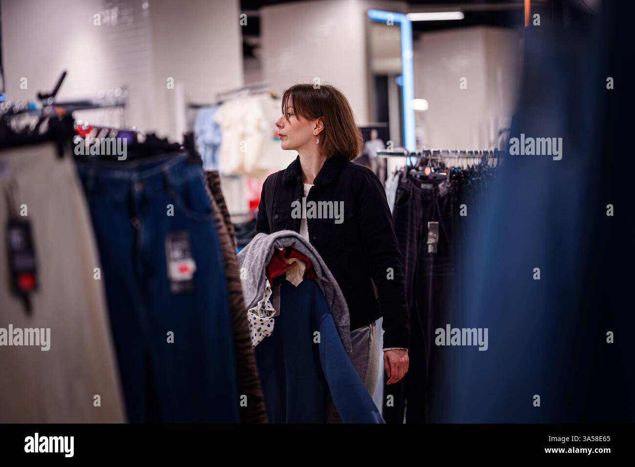 Eine Frau in einer schwarzen Jacke trägt mehrere Kleidungsstücke, während sie in einem Modegeschäft durch die Regale stöbert und Outfits für ihre Garderobe auswählt. Stockfoto