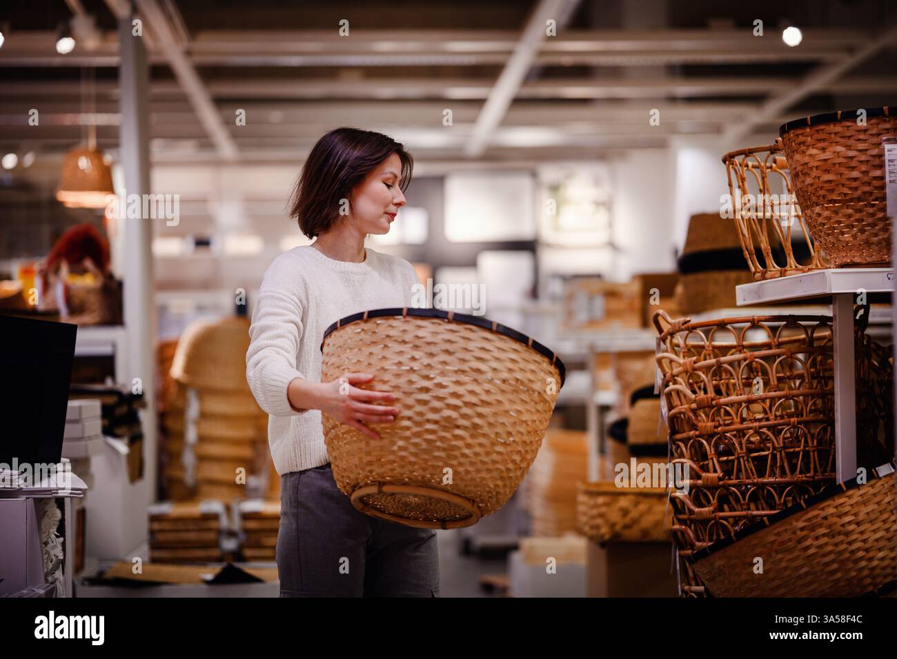 Eine Frau in einem weißen Pullover untersucht einen großen gewebten Korb in einem Heim-Geschäft und zieht ihn zum Aufbewahren oder Dekorieren beim Einkaufen von Haushaltsgegenständen in Betracht. Stockfoto