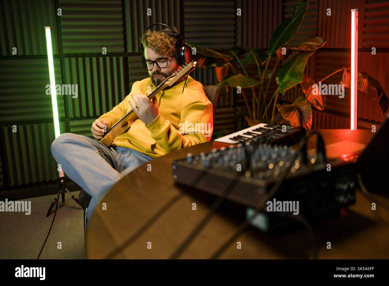 Ein fokussierter männlicher Musiker stimmt eine Gitarre vor dem Hintergrund einer digitalen Klangbearbeitung in einer zeitgenössischen Aufnahmestudioumgebung ab. Stockfoto