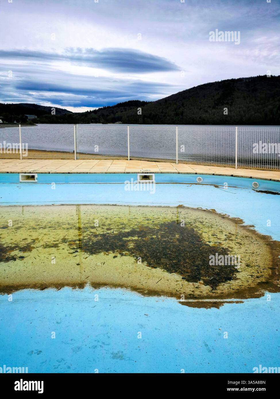 Ein vernachlässigtes Schwimmbad mit trübem Wasser steht leer, eingerahmt von einem Drahtzaun und einem ruhigen See im Hintergrund, umgeben von bewölktem Himmel - Smartphone-aufgenommenes Stockfoto