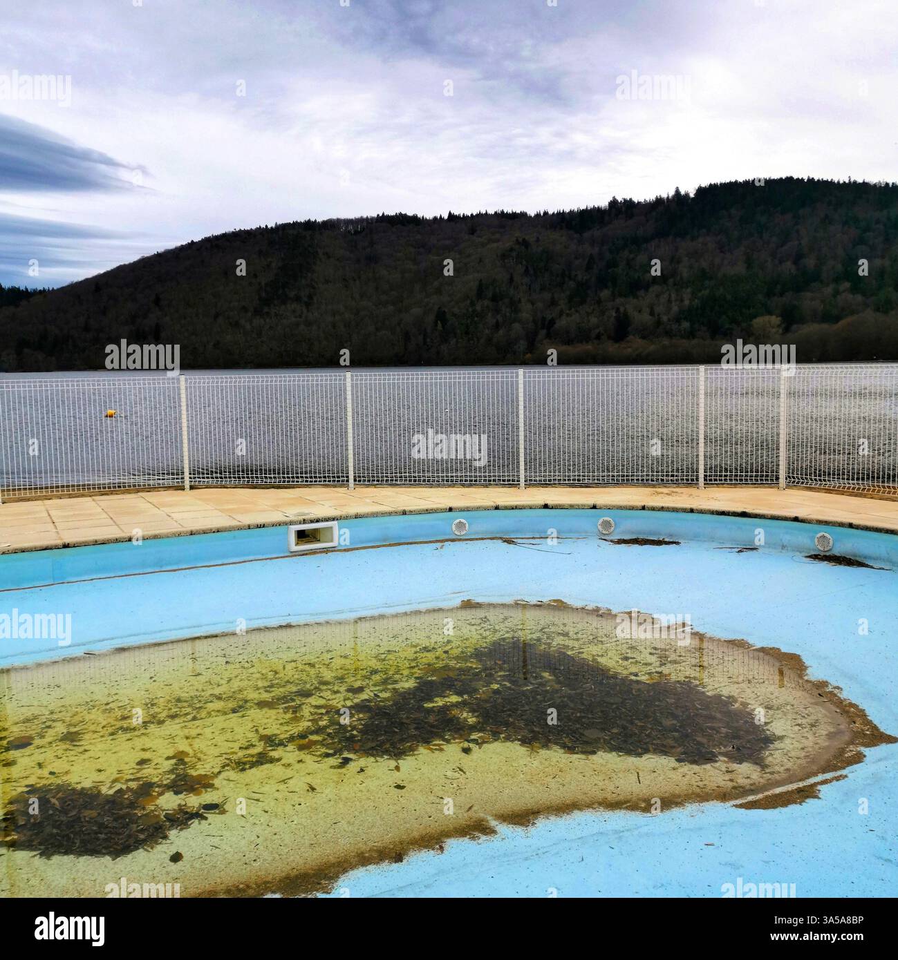 Ein vernachlässigtes Schwimmbad mit trübem Wasser steht leer, eingerahmt von einem Drahtzaun und einem ruhigen See im Hintergrund, umgeben von bewölktem Himmel - Smartphone-aufgenommenes Stockfoto