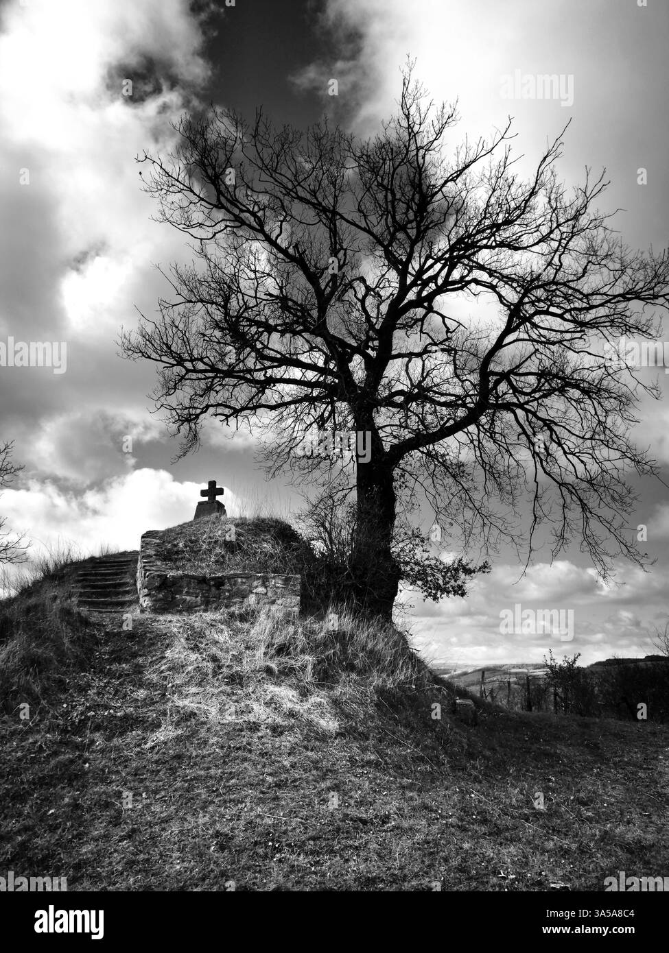 Ein einsamer Baum steht auf einem Hügel, eingerahmt von dunklen Wolken. In der Nähe ist ein Kreuz zu sehen, das auf einen Ort der Reflexion hinweist. Stockfoto