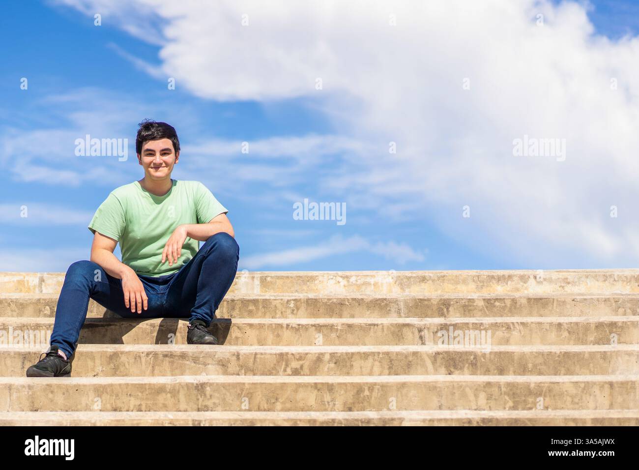 Freundlicher junger Mann, der auf Stufen sitzt und vor einem hellblauen Himmel lächelt Stockfoto