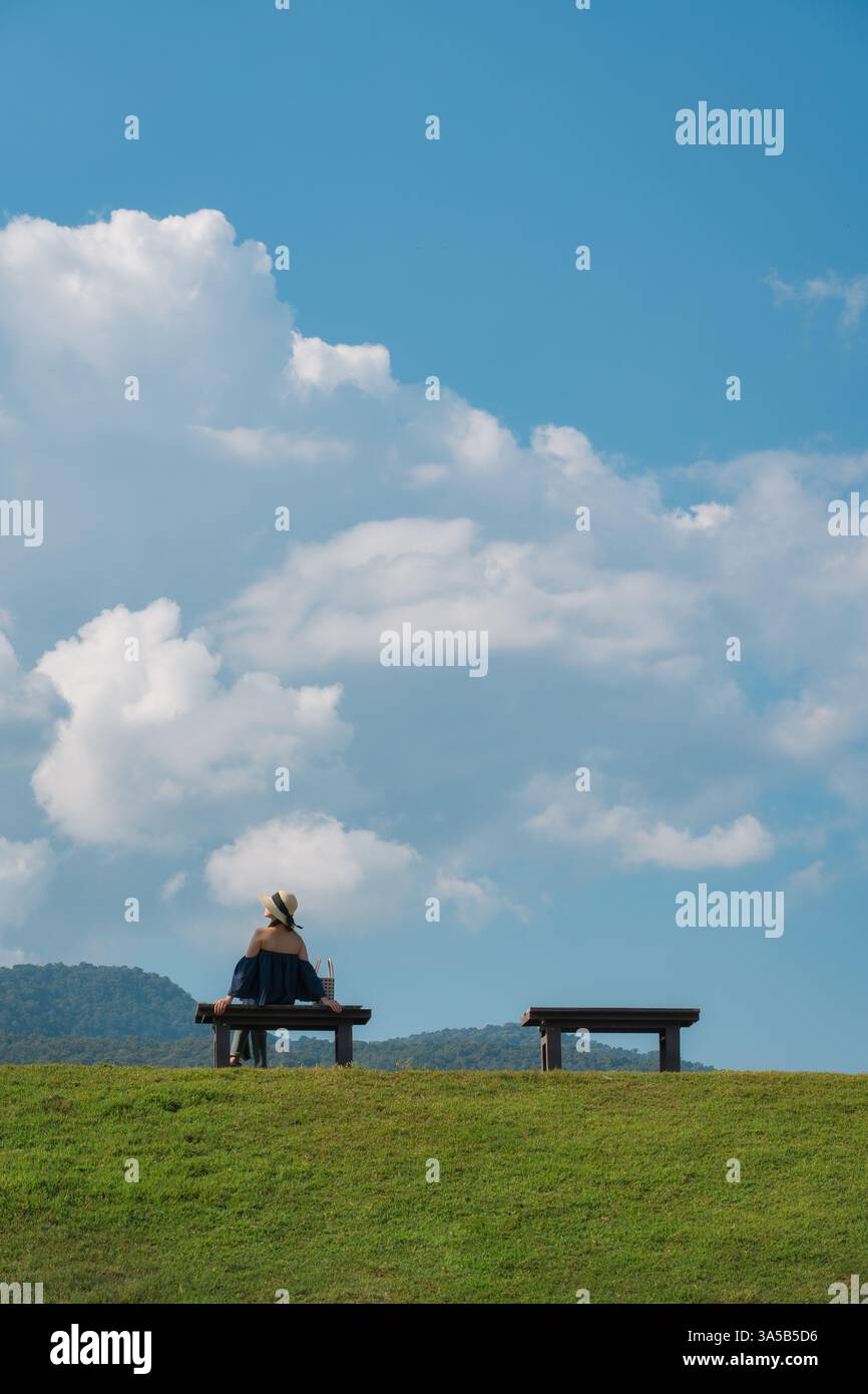 Weibliche Touristen sitzen an einer Holzbank auf einem üppigen grünen Hügel und großen flauschigen Wolken am blauen Himmel an sonnigen Tagen Stockfoto