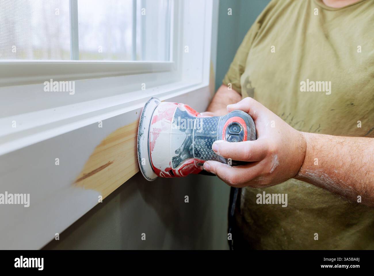Der Holzarbeiter verwendet bei Renovierungsarbeiten eine elektrische Schleifmaschine, um Holzverkleidungen am Fenster zu glätten. Stockfoto Der Holzarbeiter verwendet bei Renovierungsarbeiten eine elektrische Schleifmaschine, um Holzverkleidungen am Fenster zu glätten. Stockfoto