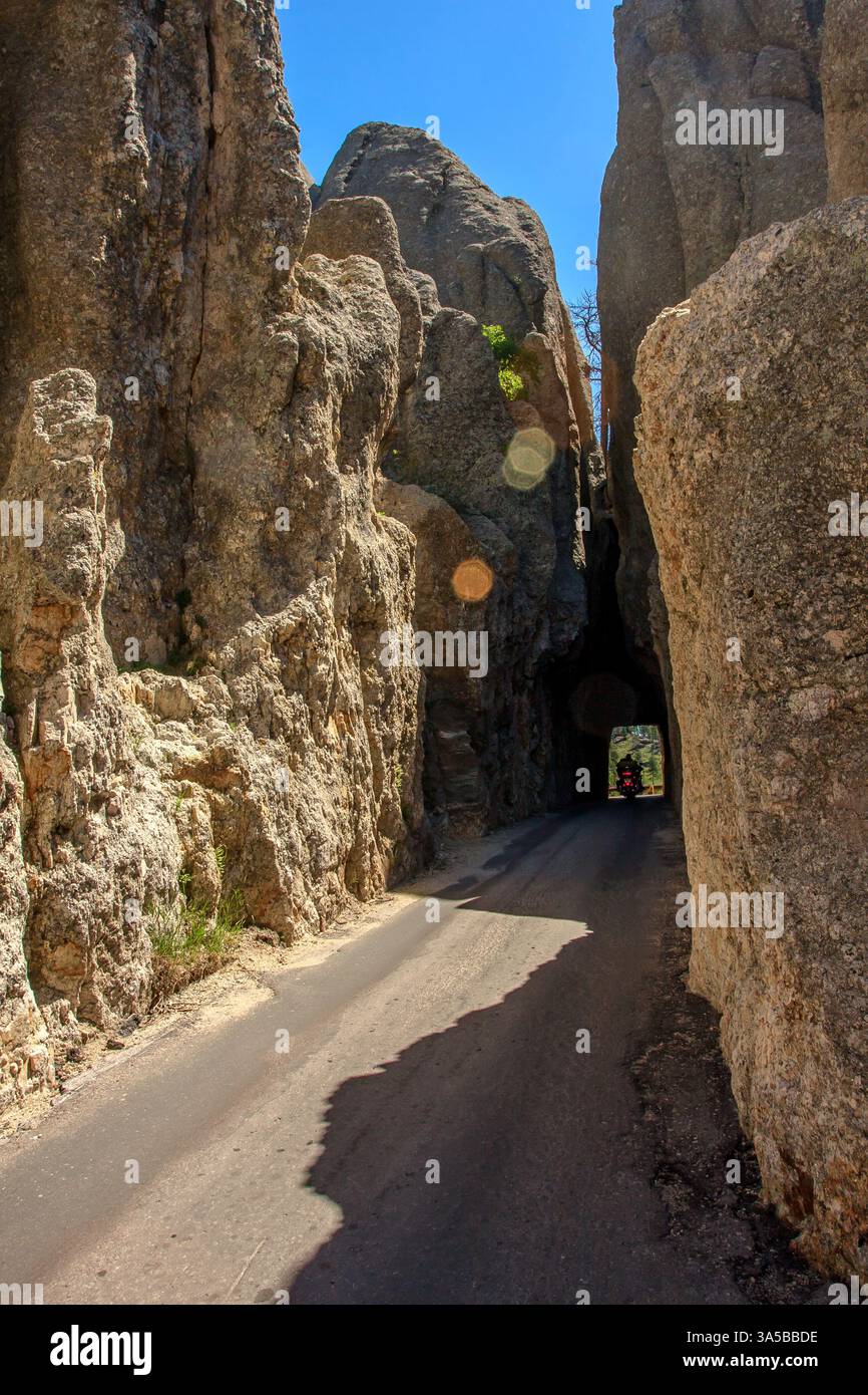 Aufnahme eines Needles Highway, der sich durch einen Tunnel schlängelt, der in hohen Felsformationen geschnitzt ist, wobei ein Motorradfahrer aus dem Tunnel unter einem c hervortritt Stockfoto
