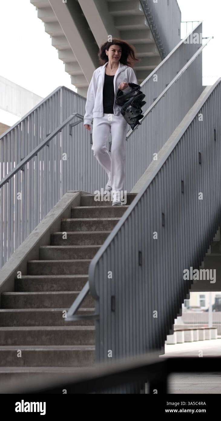 Eine Frau in einem weißen Trainingsanzug geht eine städtische Treppe hinunter und trägt Kangoo-Jumps-Rebound-Schuhe. Die strukturierte Architektur und die Activewear-Highlights Stockfoto