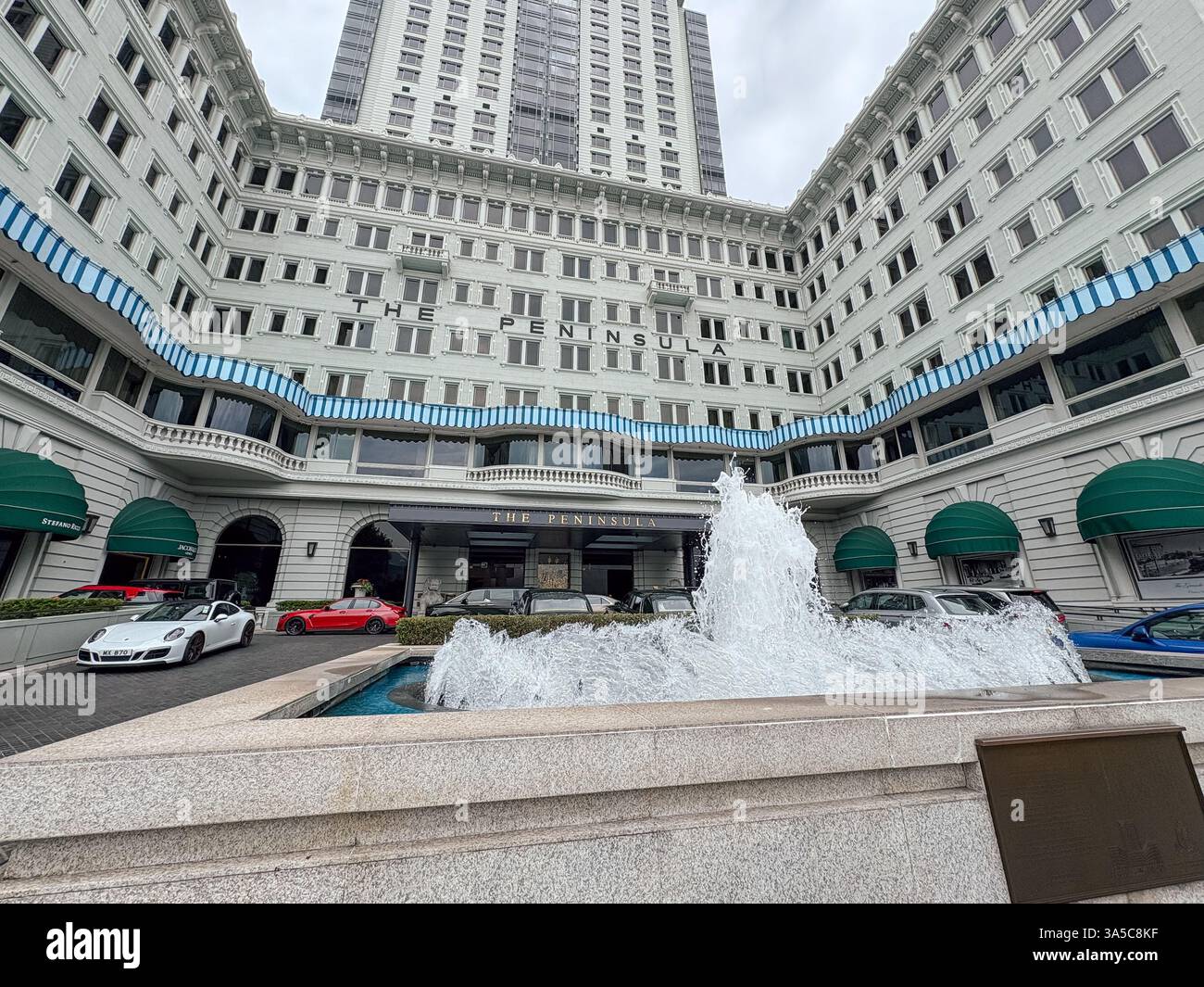 Weitwinkelblick auf das Luxushotel Peninsula Hong Kong mit Brunnen, eleganter Architektur und Autos. Stockfoto