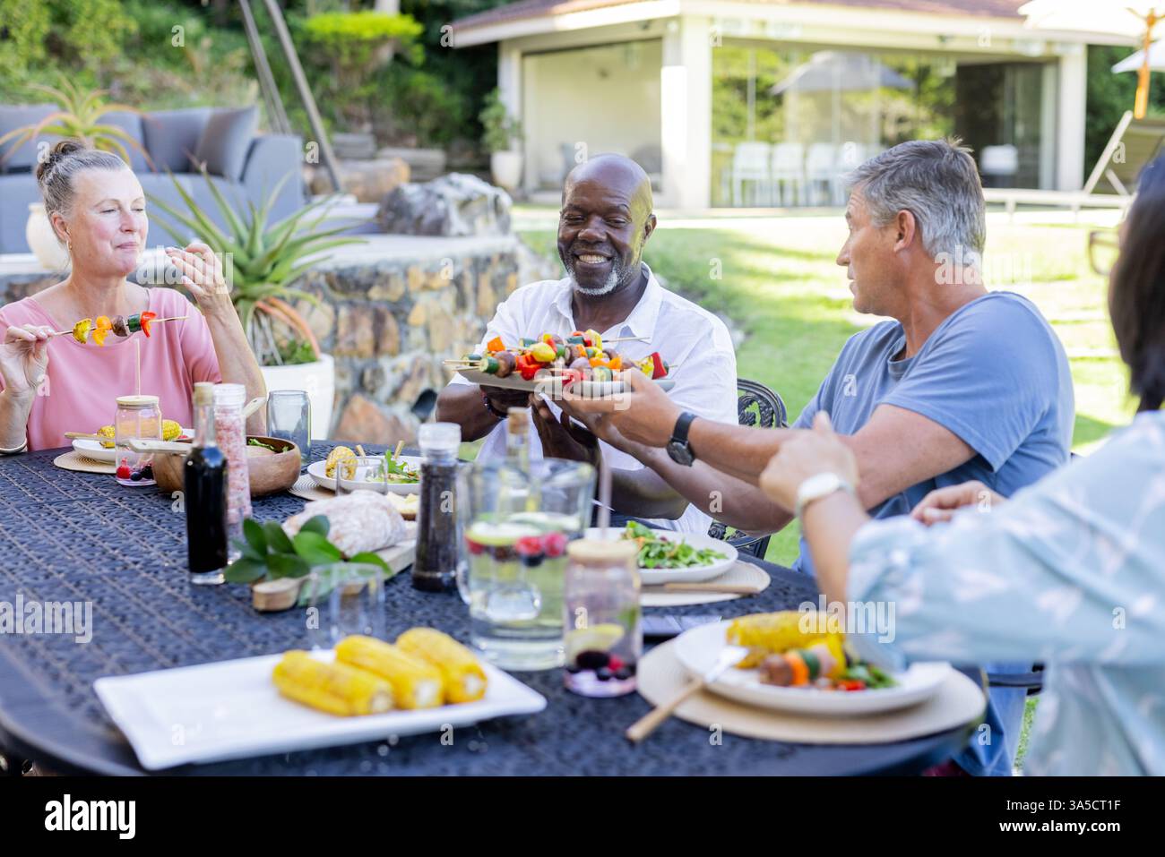 Im Garten teilen sich verschiedene Seniorenfreunde Grillgerichte und lachen gemeinsam im Freien Stockfoto