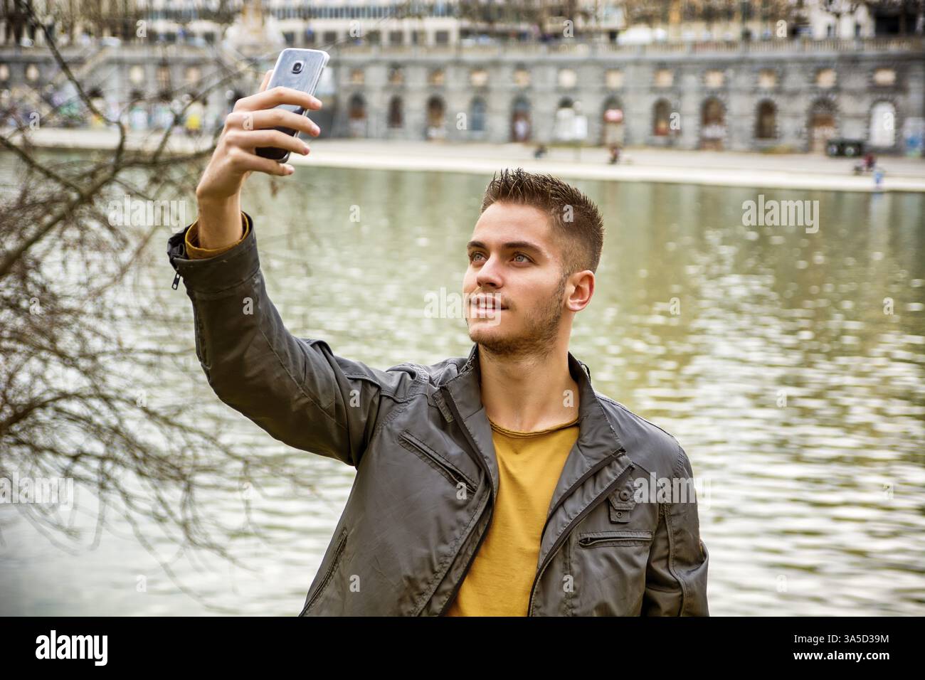 Dreiviertel Länge eines hellbraunen jungen Mannes mit grauer Jacke und Jeans, der Selfie mit seinem Smartphone vor malerisch fotografiert Stockfoto