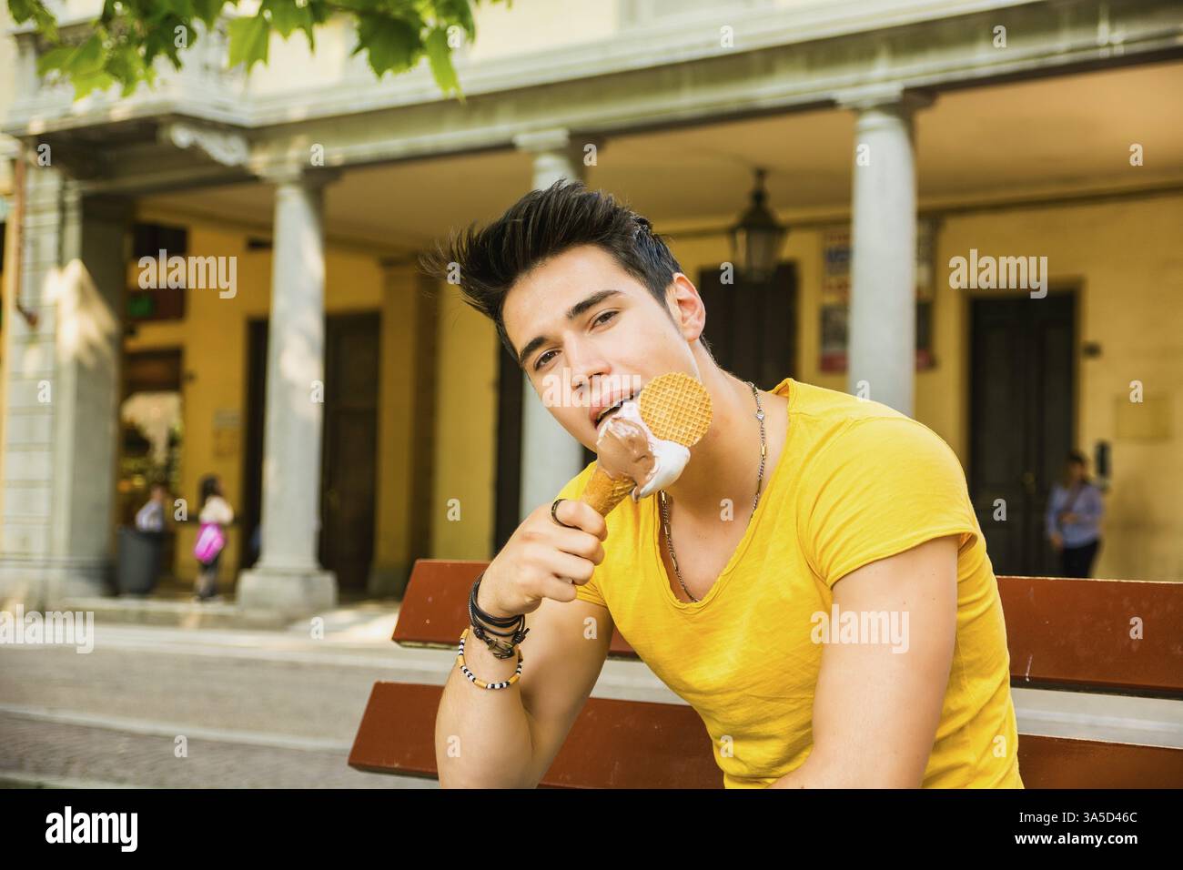 Lustiger, attraktiver junger Mann sitzt, trägt ein gelbes T-Shirt, während er draußen im Park ein leckeres Vanilleeis isst, an einem warmen Sommertag Stockfoto