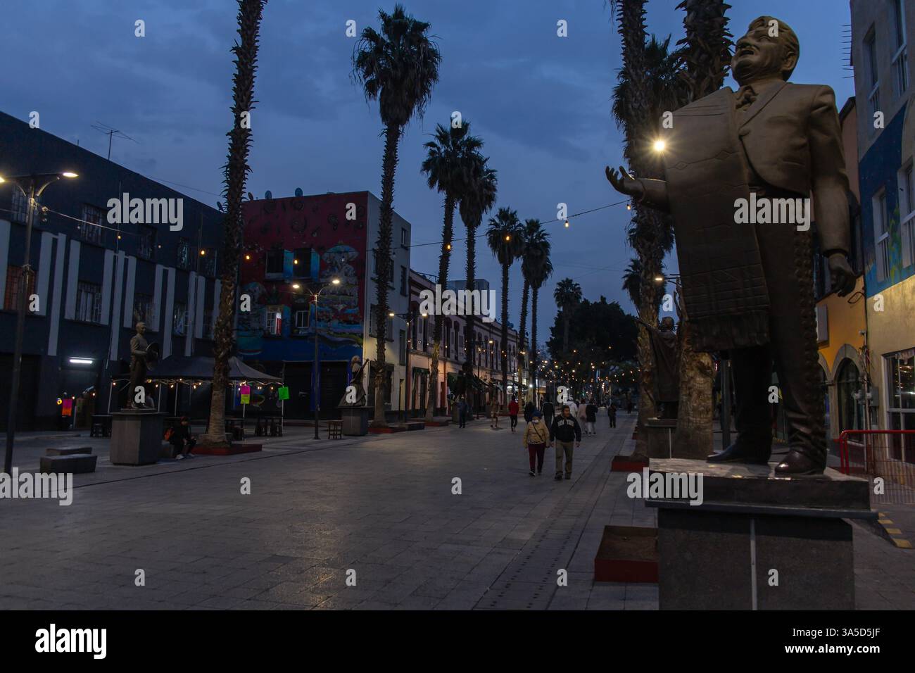 In der Abenddämmerung schlendern die Menschen entlang einer von Palmen gesäumten Fußgängerzone an der Plaza Garibaldi, umgeben von Statuen, Wandmalereien und festlichen Hängelampen. Stockfoto