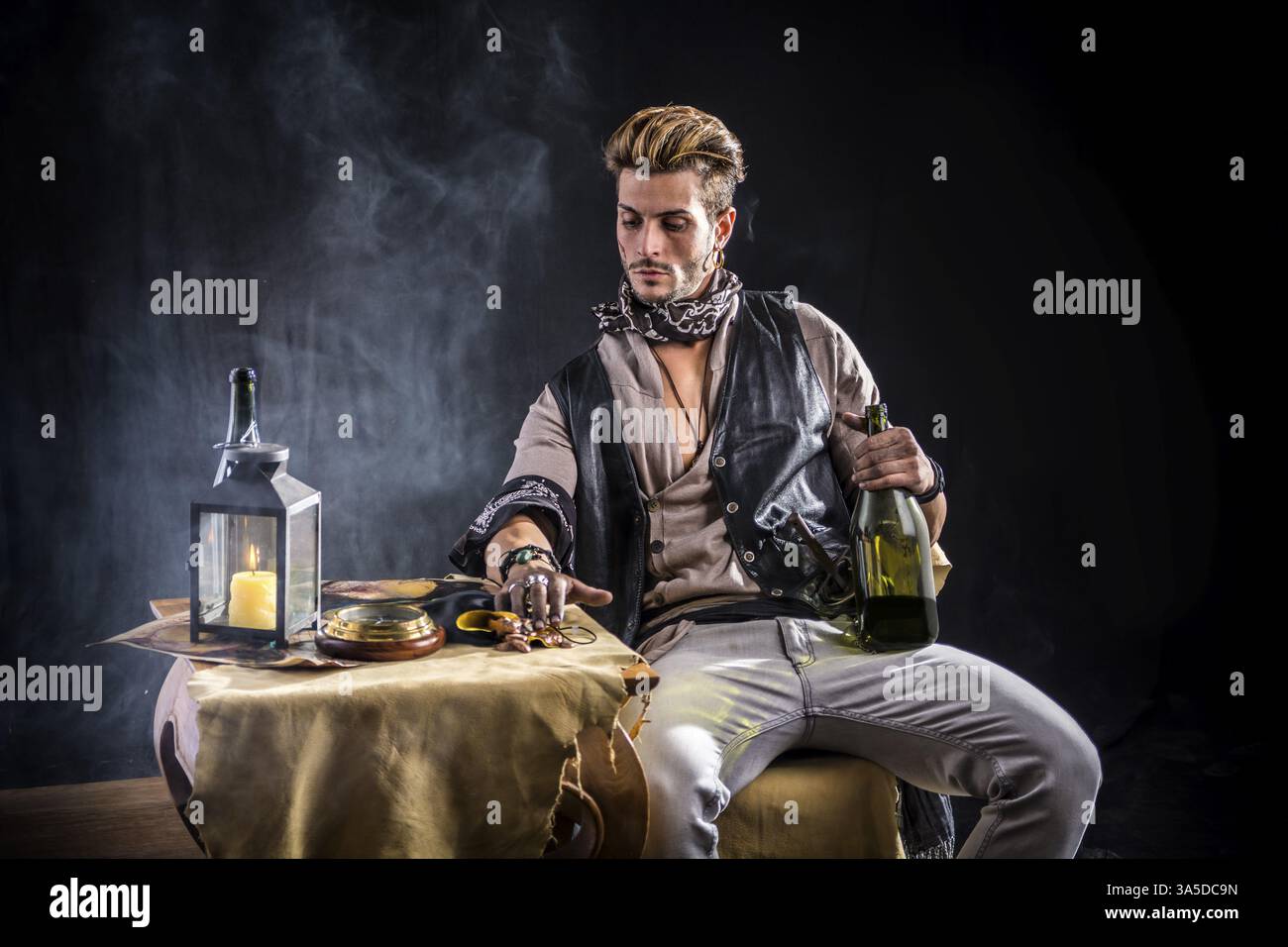 Gut aussehender junger Mann im Piratenmode-Outfit auf grauem Hintergrund. Gefangen im Studio, sitzen an einem Tisch mit einer Flasche Wein Stockfoto Gut aussehender junger Mann im Piratenmode-Outfit auf grauem Hintergrund. Gefangen im Studio, sitzen an einem Tisch mit einer Flasche Wein Stockfoto