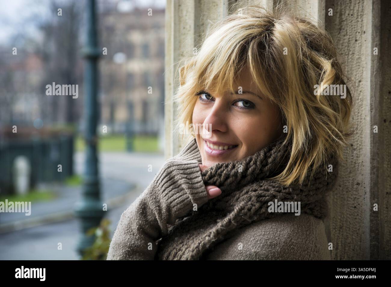 Attraktive blonde junge Frau draußen, mit Winterkleidung gegen Steinsäule, lächelnd in die Kamera Stockfoto