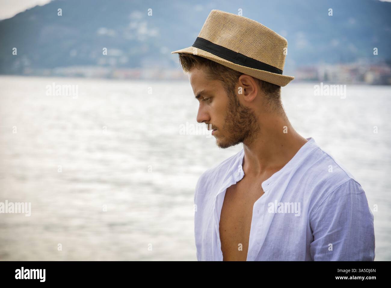 Hübscher junger Mann in trendige Kleidung, an einem Strand an einem sonnigen Sommertag, tragen ein weißes Hemd und Strohhut Stockfoto