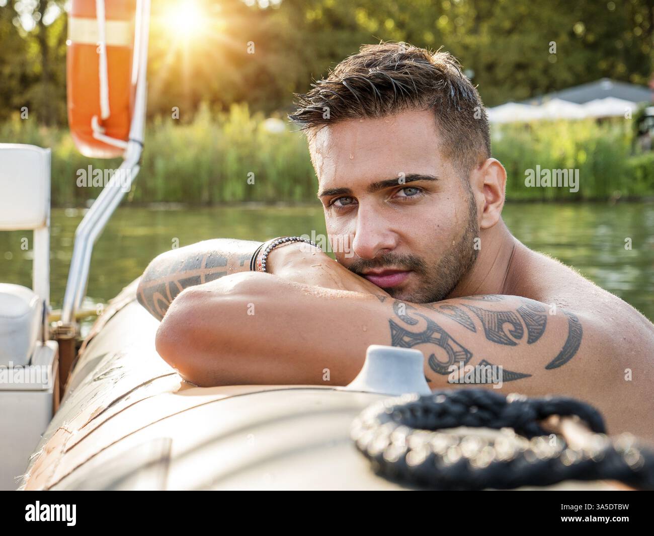 Porträt eines jungen bärtigen, gutaussehenden Mannes mit tätowiertem Körper, der sexuell in die Kamera schaut, die im Boot segelt Stockfoto