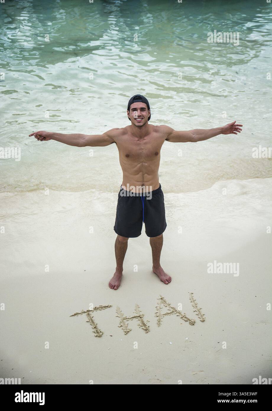 Fröhlicher junger Mann, der neben thailändischen Briefen an einem Sandstrand steht. Vertikale Außenaufnahme Stockfoto